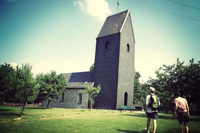 Zwei Personen gehen auf einem Weg auf eine graue Kirche mit einem schmalen Turm in einer grünen Landschaft zu.