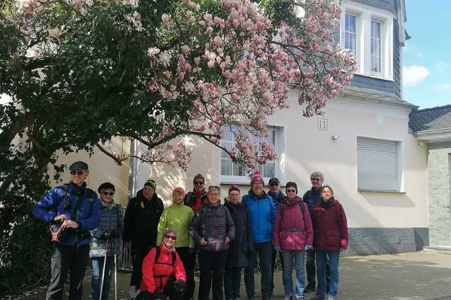 Gruppe von elf Personen posiert vor einem blühenden Magnolienbaum und einem Wohnhaus mit grauem Dach.