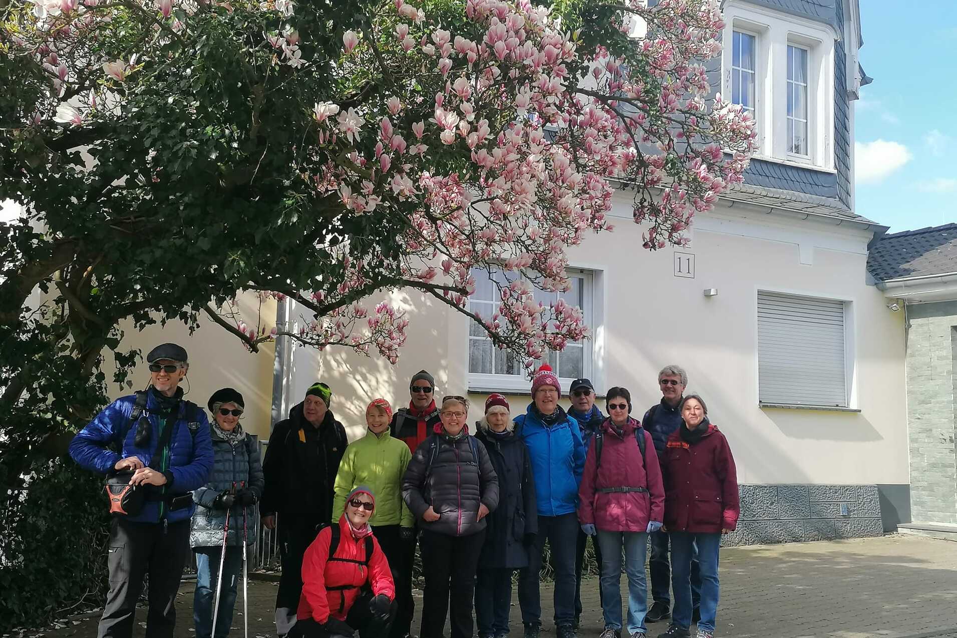 Gruppe von elf Personen posiert vor einem blühenden Magnolienbaum und einem Wohnhaus mit grauem Dach.