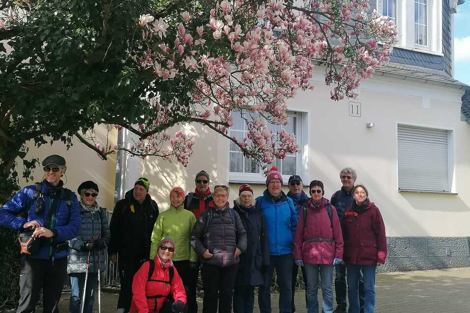 Gruppe von elf Personen posiert vor einem blühenden Magnolienbaum und einem Wohnhaus mit grauem Dach.
