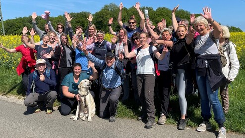 Gruppe von Menschen mit erhobenen Händen und einem Hund, posierend auf einem Weg vor blühenden Pflanzen und blauem Himmel.