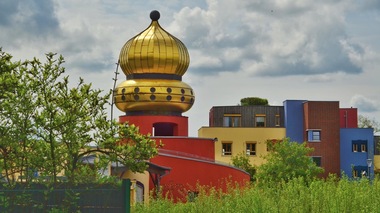 Bunte Gebäude mit goldener Kuppel vor einem bewölkten Himmel, umgeben von grünen Sträuchern.