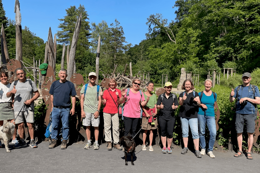 Gruppe von Wanderern mit Hunden posiert vor einer Holzskulptureninstallation im Freien an einem sonnigen Tag.