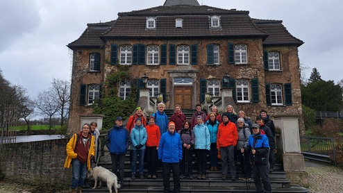 Gruppe von Menschen in bunten Jacken auf den Treppen eines alten, mehrstöckigen Hauses mit einem Hund.