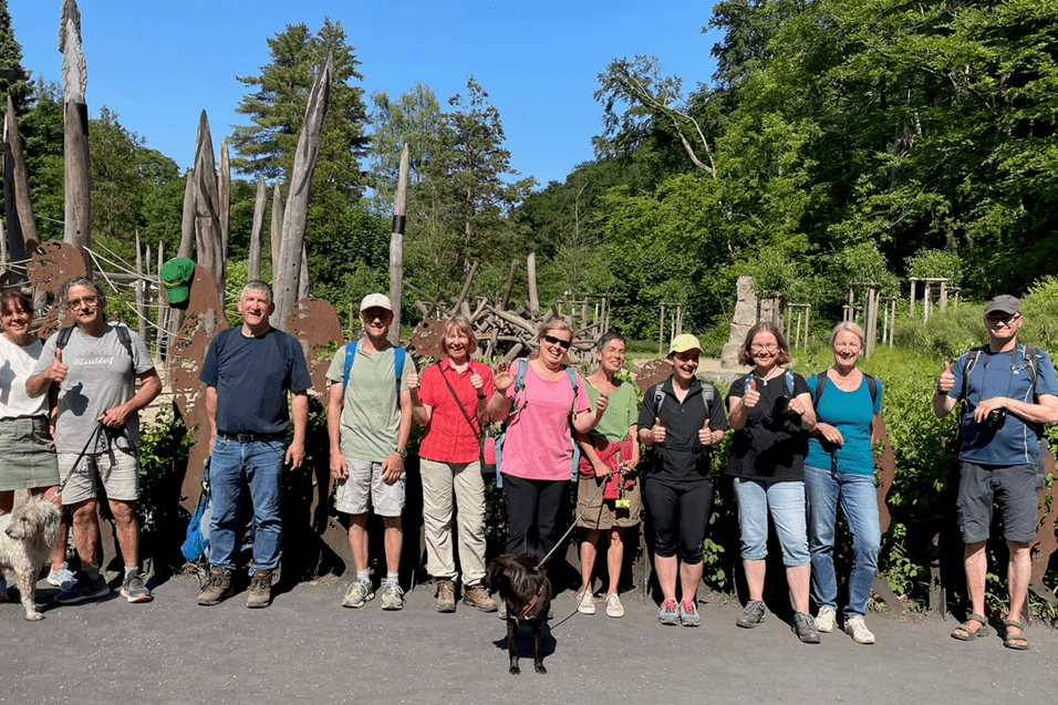 Gruppe von Wanderern mit Hunden posiert vor einer Holzskulptureninstallation im Freien an einem sonnigen Tag.