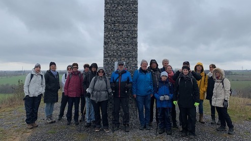 Gruppe von Menschen steht vor einem steinernen Obelisken auf einem Hügel, bewölkter Himmel im Hintergrund.