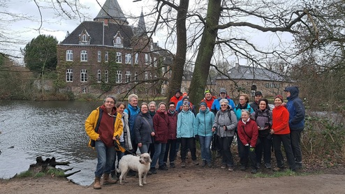 Gruppe von Wanderern in bunten Jacken posiert am Ufer eines Gewässers vor einem alten, mehrstöckigen Gebäude.