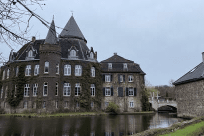 Historisches Schloss mit Türmen und einem Wassergraben, umgeben von Bäumen und einer grünen Wiese.