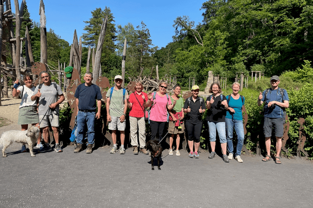 Gruppe von Wanderern mit Hunden posiert vor einer Holzskulptureninstallation im Freien an einem sonnigen Tag.