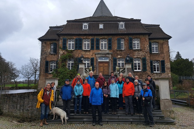 Gruppe von Menschen in bunten Jacken auf den Treppen eines alten, mehrstöckigen Hauses mit einem Hund.