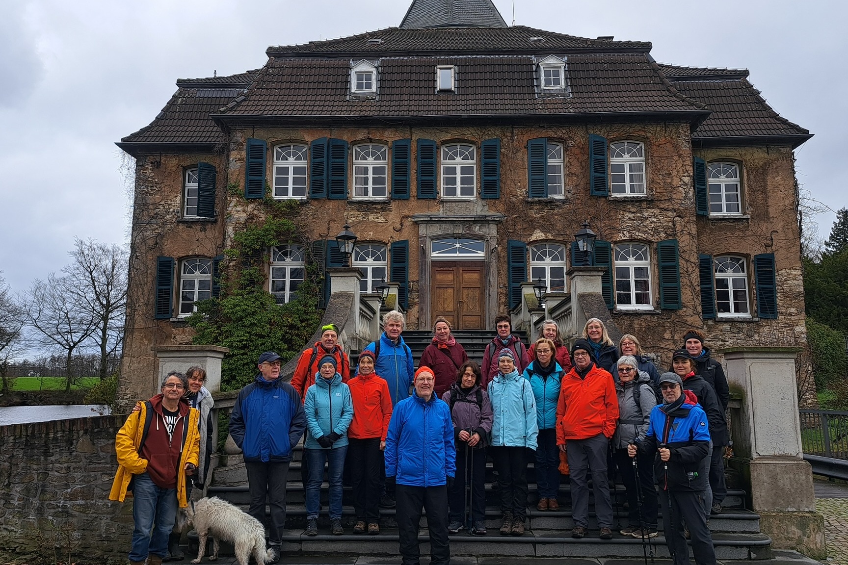 Gruppe von Menschen in bunten Jacken auf den Treppen eines alten, mehrstöckigen Hauses mit einem Hund.