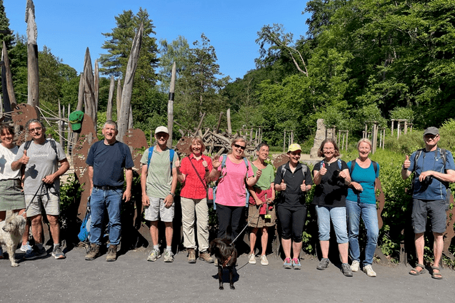 Gruppe von Wanderern mit Hunden posiert vor einer Holzskulptureninstallation im Freien an einem sonnigen Tag.