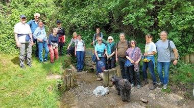 Gruppe von zwölf Personen mit zwei Hunden stehen an einer Quelle, umgeben von grünem Umland.