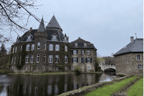 Historisches Schloss mit Türmen und einem Wassergraben, umgeben von Bäumen und einer grünen Wiese.