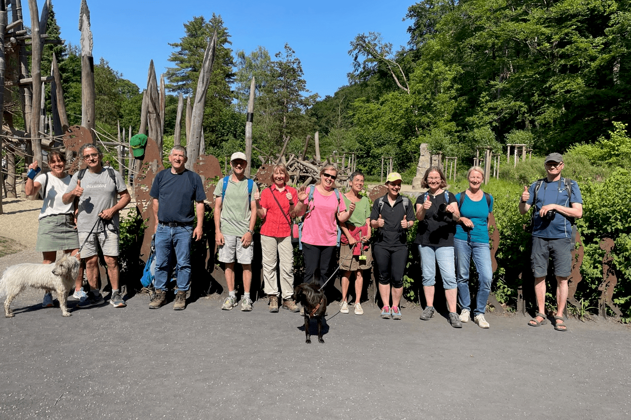 Gruppe von Wanderern mit Hunden posiert vor einer Holzskulptureninstallation im Freien an einem sonnigen Tag.