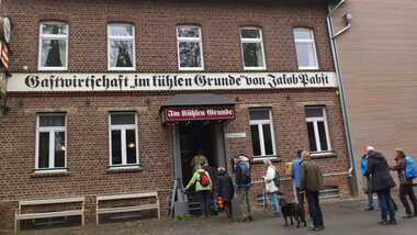 Eingang zur Gastwirtschaft "Im kühlen Grunde" mit Menschen und einem Hund vor der Fassade eines Backsteingebäudes.