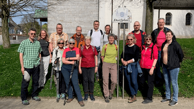 Gruppe von 16 Personen steht lächelnd vor einem Schild „Dorfplatz“ nahe einer Kirche an einem sonnigen Tag.