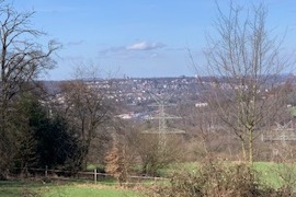 Aussicht auf eine h&uuml;gelige Landschaft mit B&auml;umen und einem kleinen Ort im Hintergrund unter einem blauen Himmel.