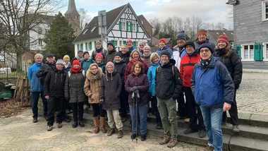 Gruppe von 30 Personen steht im Freien vor historischen Gebäuden mit einer Kirche im Hintergrund. Alle in warmer Kleidung.