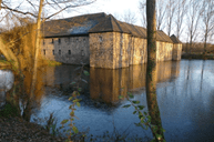 Historisches Geb&auml;ude am Ufer eines ruhigen Sees, umgeben von B&auml;umen und reflektiert im Wasser.