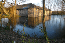 Historisches Gebäude am Ufer eines ruhigen Sees, umgeben von Bäumen und reflektiert im Wasser.