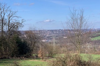 Aussicht auf eine hügelige Landschaft mit Bäumen und einem kleinen Ort im Hintergrund unter einem blauen Himmel.