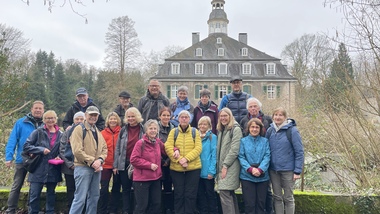 Gruppe von 20 Wanderern posiert vor einem historischen Gebäude in einer bewaldeten Umgebung an einem grauen Tag.