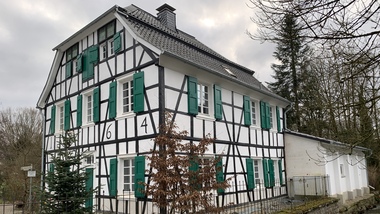 Fachwerkhaus mit weißer Fassade und grünen Fensterläden, umgeben von Bäumen und einem kleinen Gewässer.