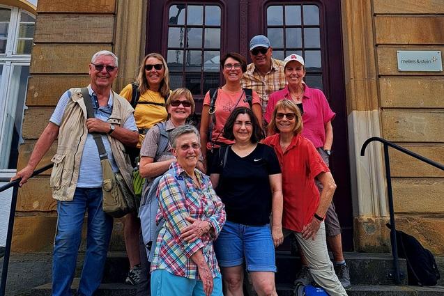 Gruppe von neun Personen auf Stufen vor einem historischen Geb&auml;ude, alle in Freizeitkleidung und l&auml;chelnd.