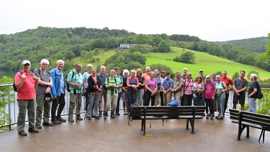 Gruppentreffen von Wanderern auf Aussichtspunkt mit Blick auf grüne Hügel und See im Hintergrund.