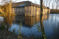 Historisches Geb&auml;ude am Ufer eines ruhigen Sees, umgeben von B&auml;umen und reflektiert im Wasser.