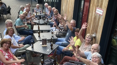 Gruppenfoto von 20 Personen, die an Tischen im Freien sitzen und in die Kamera lachen und winken.