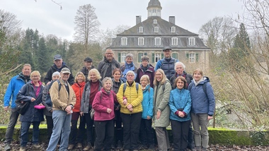 Gruppe von 20 Wanderern posiert vor einem historischen Gebäude in einer bewaldeten Umgebung an einem grauen Tag.