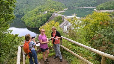 Drei Frauen stehen am Ufer eines Stausees, umgeben von Grün, Blick auf staumauer im Hintergrund.