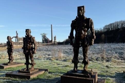 Vier metallene Skulpturen stehen in einem frostigen Feld, im Hintergrund sind Industrieanlagen und ein blauer Himmel sichtbar.