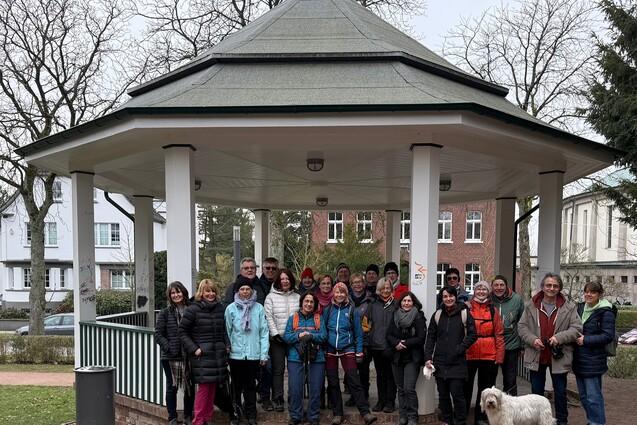 Gruppe von Personen in Winterkleidung steht unter einem Pavillon in einem Park, ein Hund sitzt vorne.