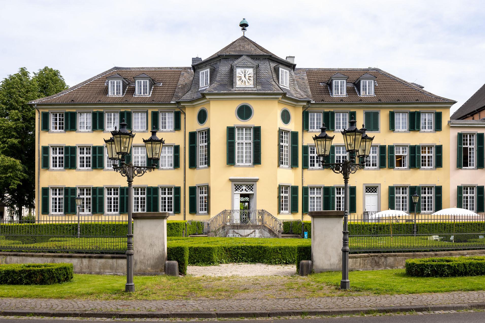 Historisches gelbes Geb&auml;ude mit gr&uuml;nen Fensterl&auml;den und zwei Stra&szlig;enlaternen im Vordergrund, umgeben von B&auml;umen.