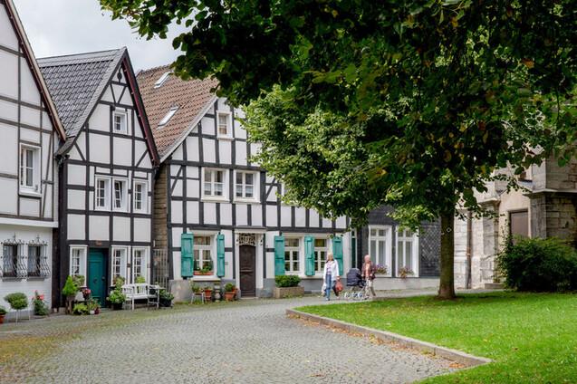 Fachwerkhäuser in einer ruhigen Gasse, umgeben von Bäumen und rasen, mit Passanten auf dem Weg.