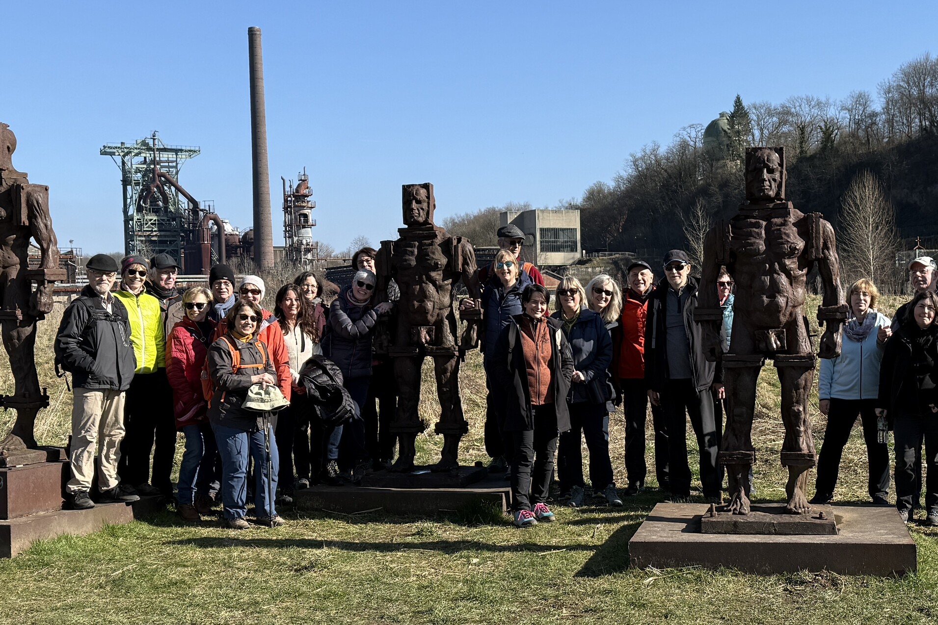Gruppe von Menschen posiert vor mehreren gro&szlig;en, rostigen Skulpturen in einem Industriegebiet an einem klaren Himmel.