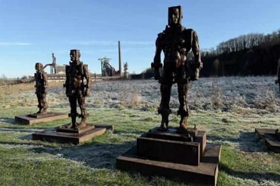 Vier metallene Skulpturen stehen in einem frostigen Feld, im Hintergrund sind Industrieanlagen und ein blauer Himmel sichtbar.