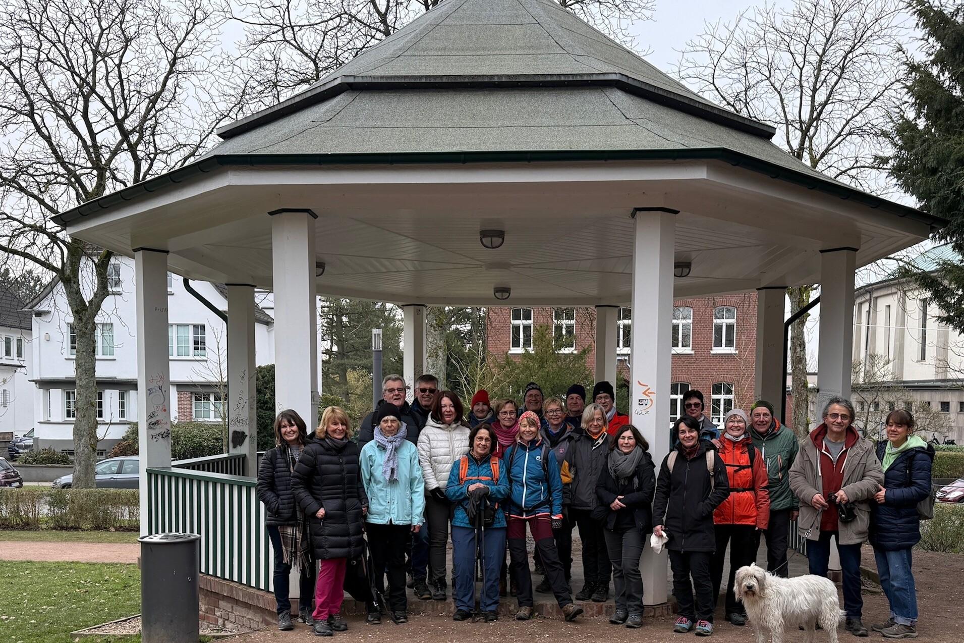 Gruppe von Personen in Winterkleidung steht unter einem Pavillon in einem Park, ein Hund sitzt vorne.