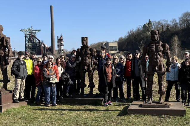 Gruppe von Menschen posiert vor mehreren großen, rostigen Skulpturen in einem Industriegebiet an einem klaren Himmel.