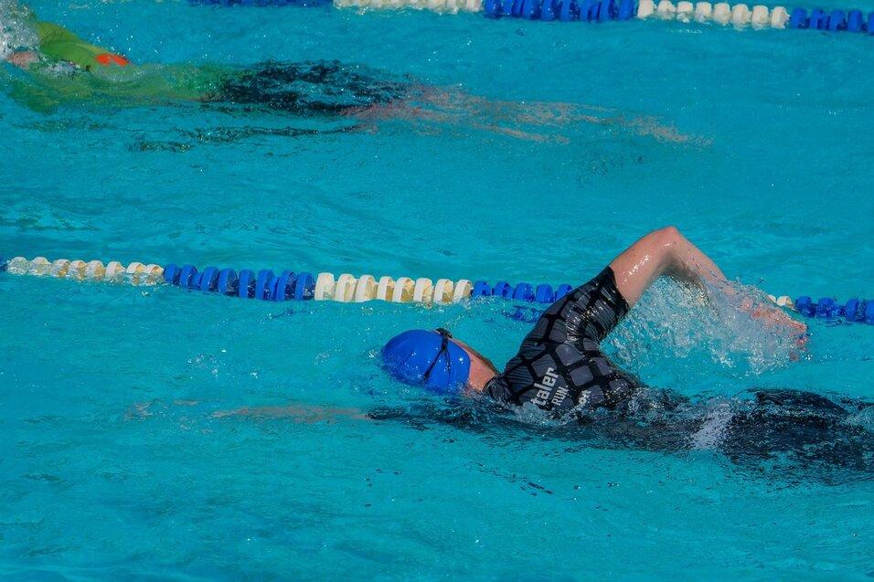 Schwimmer in blauer Badekappe und schwarzem Anzug beim Kraulen in einem Schwimmbecken mit blauem Wasser.