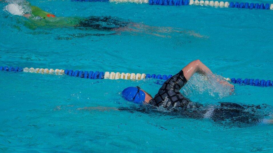 Schwimmer in blauer Badekappe und schwarzem Anzug beim Kraulen in einem Schwimmbecken mit blauem Wasser.
