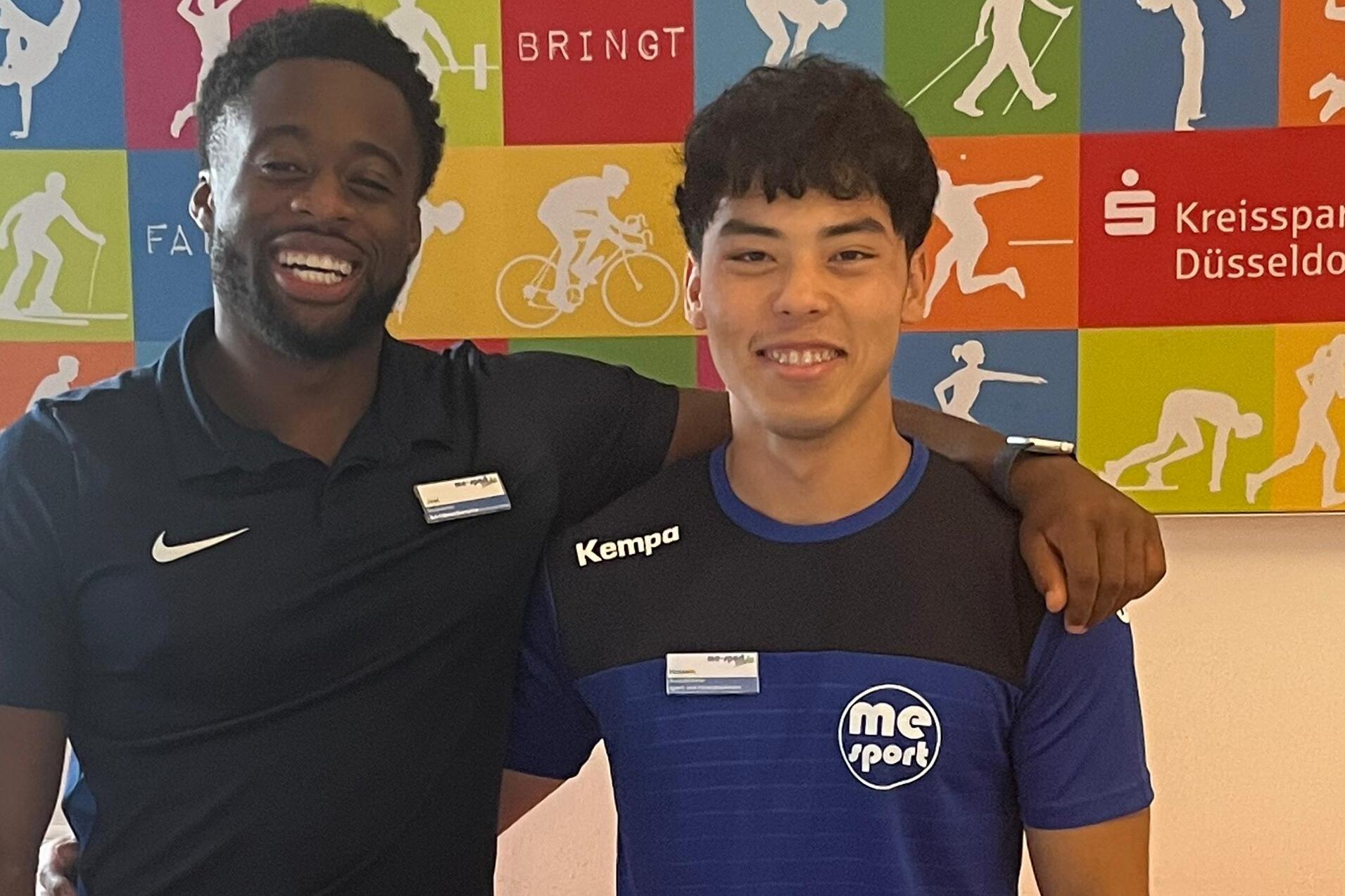 Zwei junge Männer stehen lächelnd vor einer farbigen Wand mit sportlichen Motiven und einem orangefarbenen Tisch.