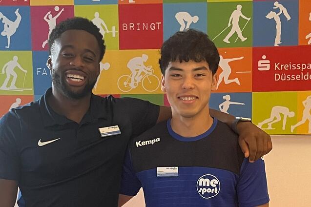 Zwei junge Männer stehen lächelnd vor einer farbigen Wand mit sportlichen Motiven und einem orangefarbenen Tisch.