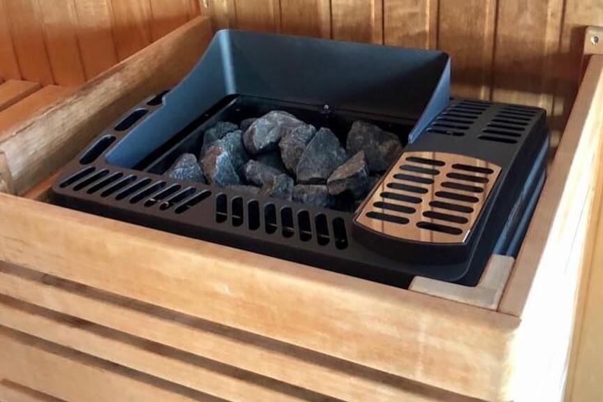 Saunasteuerung mit schwarzen Steinen in einem eckigen Holzbecken, umgeben von einer Holzvertäfelung.
