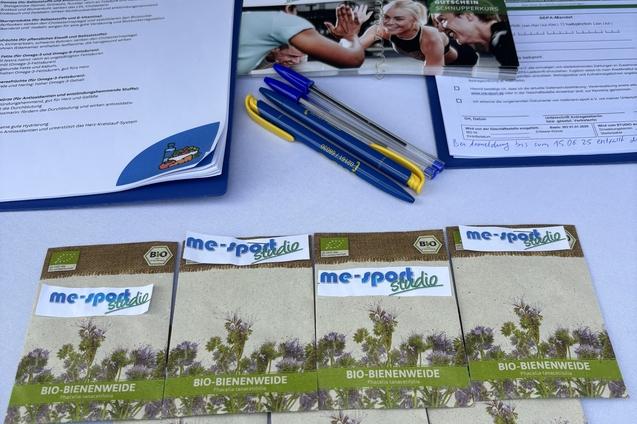 Tisch mit Informationsmaterialien, darunter Einkaufslisten, Gutscheine und Broschüren zur Bienenweide im Vordergrund.