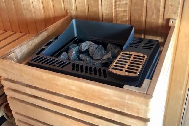 Saunasteuerung mit schwarzen Steinen in einem eckigen Holzbecken, umgeben von einer Holzvertäfelung.