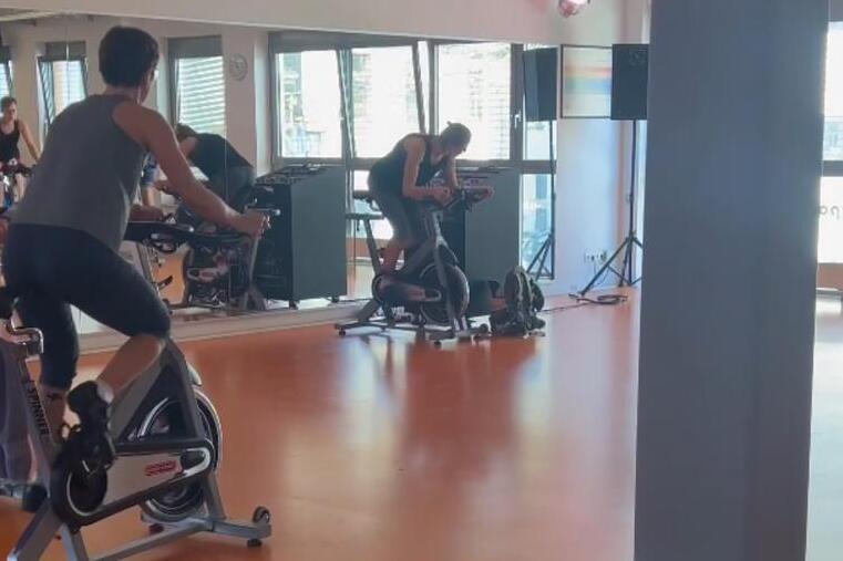 Menschen sitzen auf Fahrr&auml;dern in einem Fitnessraum mit orangefarbenem Boden und gro&szlig;en Fenstern.